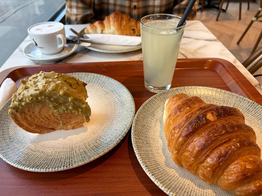 Croissant, Pistazien-Croissant, Cappuccino und ein Glas Limonade auf einem Tablett